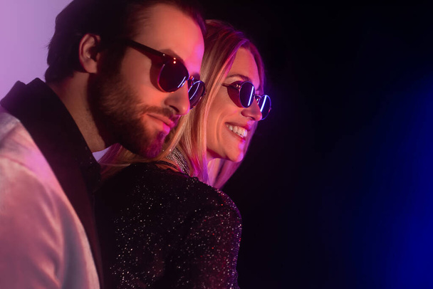 Cheerful blonde woman in sunglasses standing near bearded boyfriend on purple and black background  - Fotografie, Obrázek