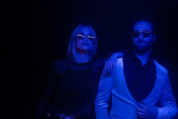 Fashionable couple in sunglasses standing isolated on black with blue lighting  - Fotografie, Obrázek