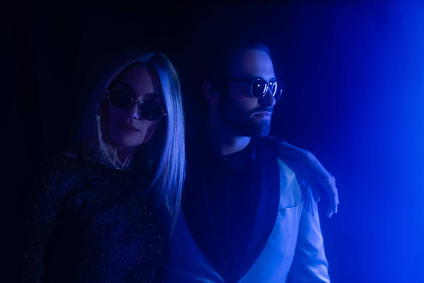 Blonde woman in sunglasses hugging stylish boyfriend on black with blue lighting  - Fotoğraf, Görsel