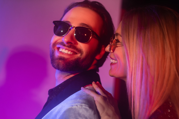 Smiling blonde woman hugging boyfriend in sunglasses on purple background  - Foto, afbeelding