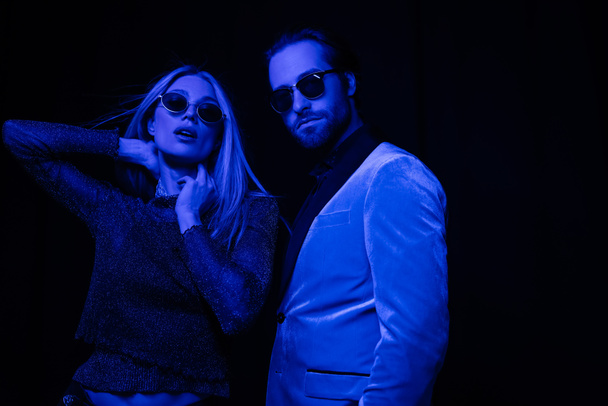 Stylish woman touching neck near boyfriend in sunglasses isolated on black with blue lighting  - Photo, Image
