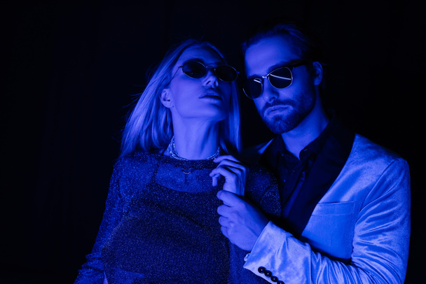 Young man in sunglasses touching hand of blonde girlfriend during party isolated on black with blue lighting  - Фото, изображение