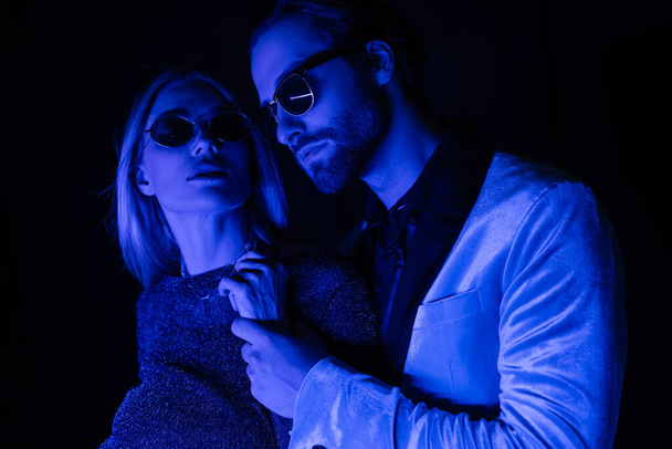 Stylish young man touching hand of girlfriend in sunglasses isolated on black with blue lighting  - Фото, изображение