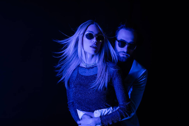 Man in sunglasses hugging blonde girlfriend during party isolated on black with blue lighting  - Foto, Bild