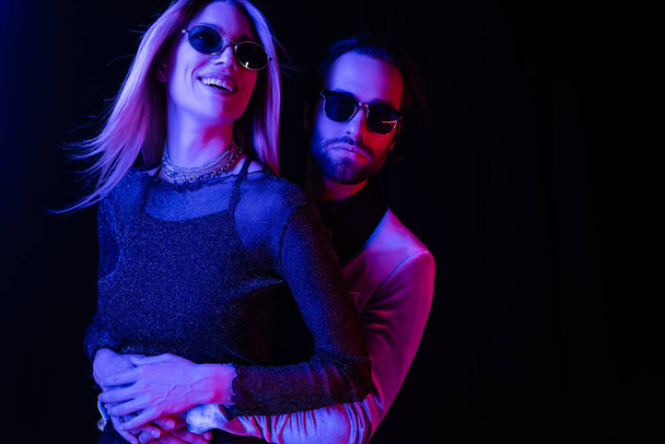 Young man in jacket hugging smiling girlfriend in sunglasses isolated on black with blue lighting  - Fotó, kép