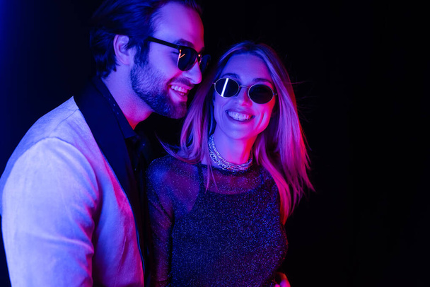 Smiling blonde woman standing near boyfriend in sunglasses isolated on black with colorful lighting  - Foto, Imagem