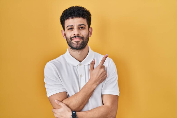 Uomo arabo in piedi su sfondo giallo che punta con il dito della mano verso il lato mostrando pubblicità, viso serio e calmo  - Foto, immagini
