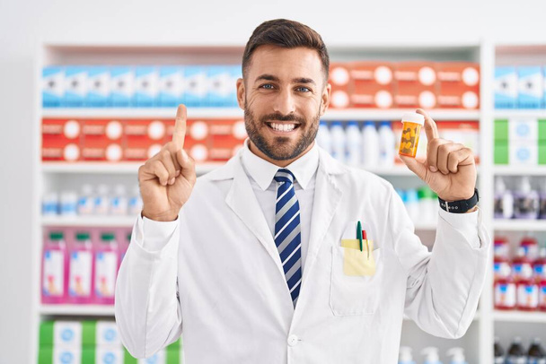 Красивый латиноамериканец, работающий в аптеке, держит таблетки, улыбаясь с идеей или вопросом, указывая пальцем со счастливым лицом, номер один  - Фото, изображение