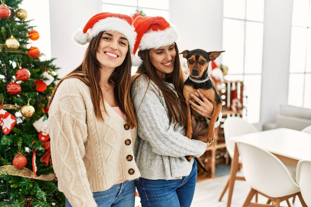 Dos mujeres sonriendo seguro abrazando perro de pie junto al árbol de Navidad en casa - Foto, imagen