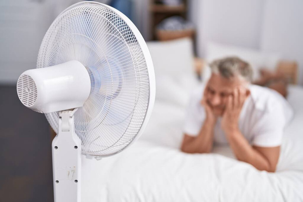 Hombre de pelo gris de mediana edad usando ventilador acostado en la cama en el dormitorio - Foto, imagen