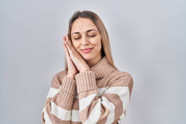 Young blonde woman wearing turtleneck sweater over isolated background sleeping tired dreaming and posing with hands together while smiling with closed eyes.  - 写真・画像