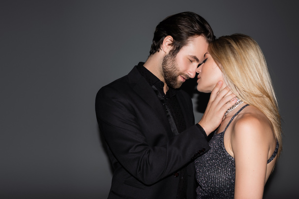 Side view of bearded man in jacket touching blonde girlfriend on grey background  - Zdjęcie, obraz