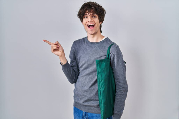 Újrahasznosítható táskát viselő fiatalember, mosolyogva, boldogan mutogatva, kezével és ujjával oldalra.  - Fotó, kép