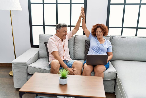 Мужчина и женщина среднего возраста используют ноутбук пять поднятых рук сидя на диване дома - Фото, изображение