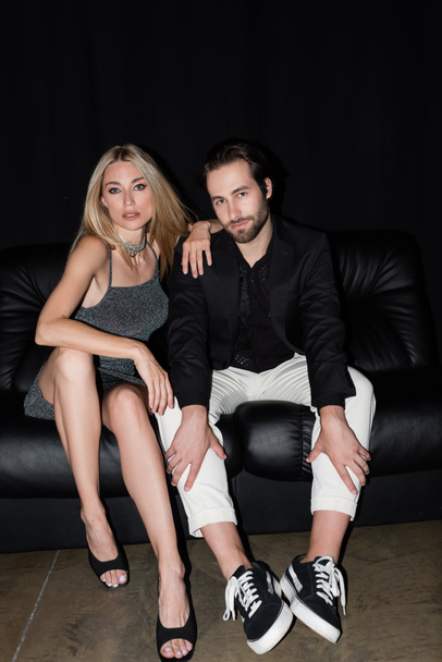 Blonde woman in dress looking at camera near boyfriend on leather couch on black background  - Foto, Imagen