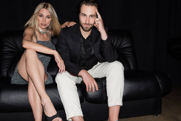 Stylish couple looking at camera while sitting on leather couch on black background  - Foto, immagini