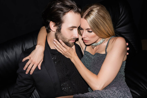 Sexy blonde woman in dress touching bearded man on leather couch isolated on black  - Photo, image