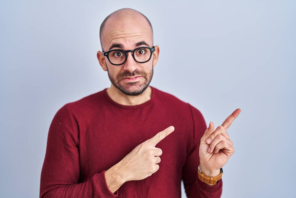 Fiatal, szakállas, fehér háttér fölött álló kopasz férfi szemüvegben, aki mindkét kezével nyugtalan és ideges, aggódó és meglepett arckifejezéssel  - Fotó, kép