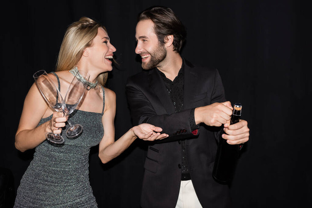 Side view of positive blonde woman in dress holding glasses near boyfriend with champagne isolated on black  - Φωτογραφία, εικόνα