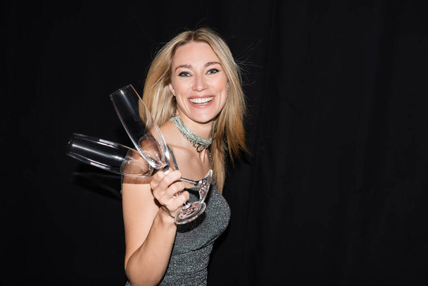 Smiling blonde woman in dress holding champagne glasses and looking at camera isolated on black  - Foto, Bild
