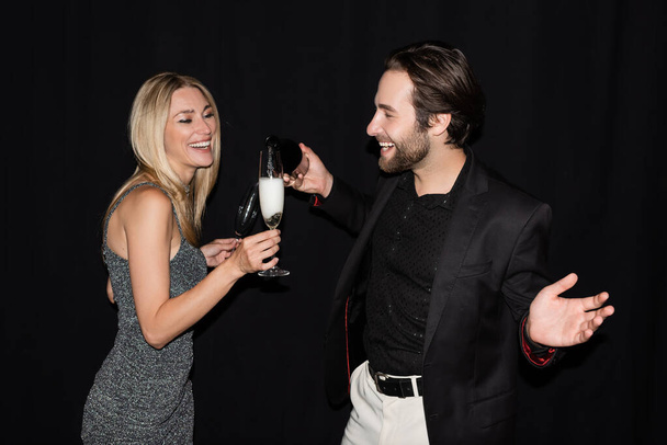 Positive man in jacket pouring champagne near blonde girlfriend isolated on black  - Фото, зображення