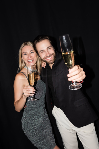 Positive couple looking at camera while holding glasses of champagne isolated on black  - Valokuva, kuva