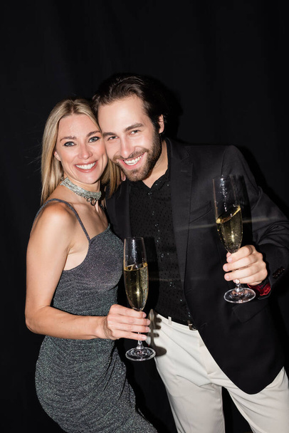 Cheerful man holding glass of champagne near stylish blonde girlfriend isolated on black  - Photo, image