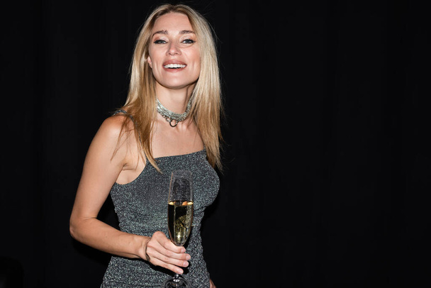 Sexy blonde woman in dress holding champagne and smiling at camera isolated on black  - Foto, Imagem