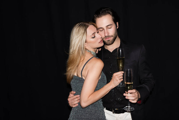 Sexy woman in dress holding glass of champagne and hugging boyfriend isolated on black  - Fotó, kép