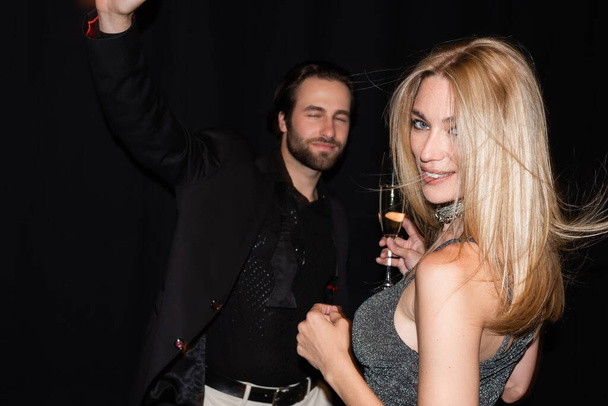 Smiling blonde woman holding glass of champagne while boyfriend dancing isolated on black  - Fotó, kép
