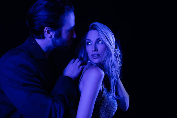 Young man touching seductive blonde girlfriend with blue lighting isolated on black  - Foto, afbeelding