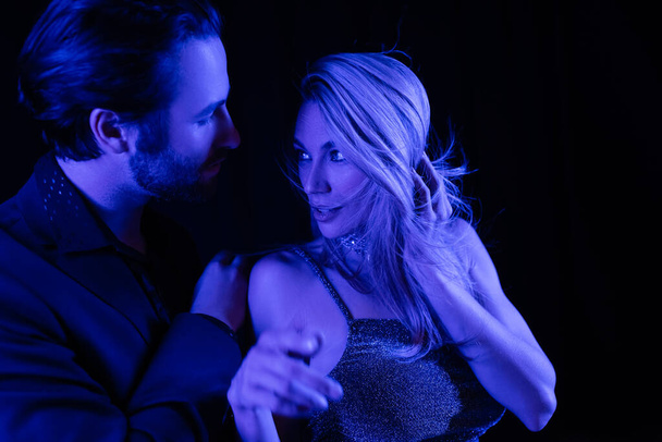 Young man touching blonde girlfriend with blue lighting isolated on black  - Valokuva, kuva