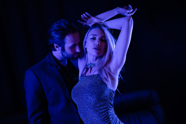 Passionate man standing near sensual girlfriend in dress with blue lighting on black background  - 写真・画像