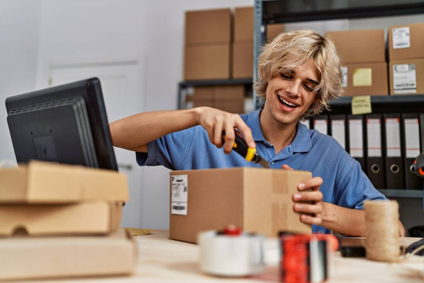 Молодой блондин, рабочий электронной коммерции, распаковывает картонную коробку в офисе - Фото, изображение