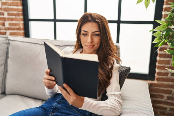 Young hispanic woman reading book sitting on sofa at home - Foto, Imagem