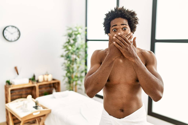 Jeune homme afro-américain torse nu portant une serviette debout au centre de beauté choqué couvrant la bouche avec les mains par erreur. concept secret.  - Photo, image