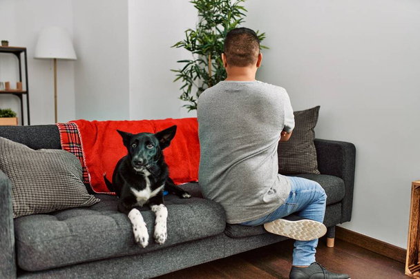 Young latin man and dog sitting on the sofa at home standing backwards looking away with crossed arms  - Foto, Imagen
