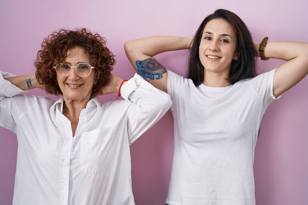 Mère et fille hispaniques portant un t-shirt blanc décontracté sur fond rose relaxant et étirant, bras et mains derrière la tête et le cou souriant heureux  - Photo, image