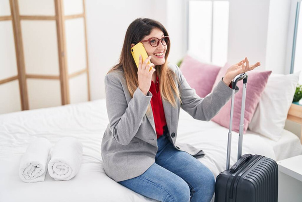 Молода іспаномовна жінка бізнесмен говорить на смартфоні, сидячи на ліжку в готельному номері
 - Фото, зображення
