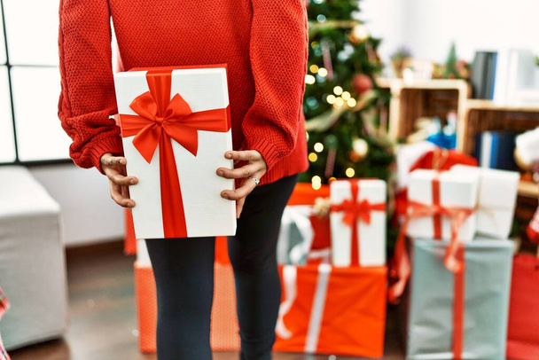 Young hispanic woman holding gift on back standing by christmas tree at home - Foto, afbeelding