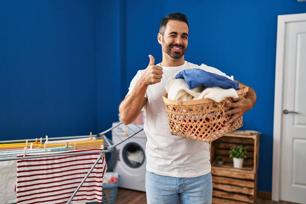 Junger hispanischer Mann mit Bart, Wäschekorb haltend, glücklich und positiv lächelnd, Daumen hoch und Zustimmungszeichen  - Foto, Bild