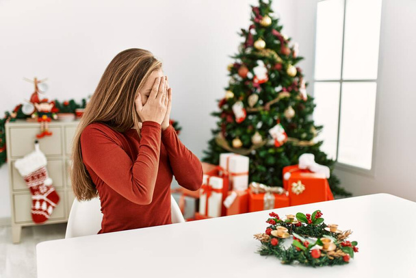 Kaukasische junge blonde Frau, die auf dem Tisch am Weihnachtsbaum sitzt, mit traurigem Gesichtsausdruck, der das Gesicht mit den Händen bedeckt, während sie weint. Depressionskonzept.  - Foto, Bild