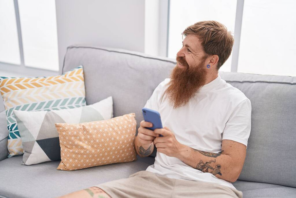 Young redhead man using smartphone sitting on sofa at home - Φωτογραφία, εικόνα