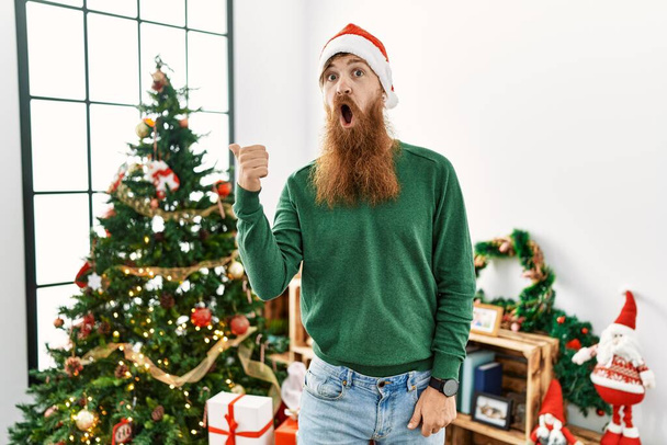 Uomo dai capelli rossi con la barba lunga che indossa il cappello di Natale dall'albero di Natale sorpreso indicando con il dito della mano di lato, bocca aperta espressione stupita.  - Foto, immagini