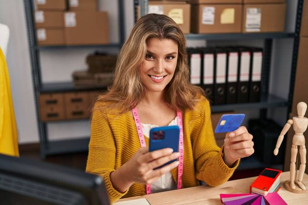 Vestidor de mujer rubia joven usando teléfono inteligente y tarjeta de crédito en el estudio de diseño - Foto, imagen