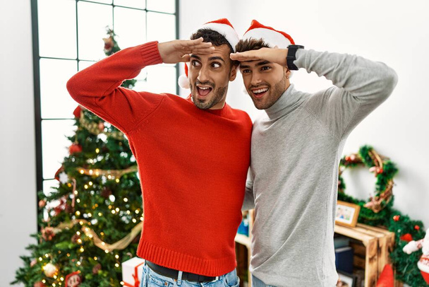 Young gay couple standing by christmas tree wearing hat very happy and smiling looking far away with hand over head. searching concept.  - Photo, Image