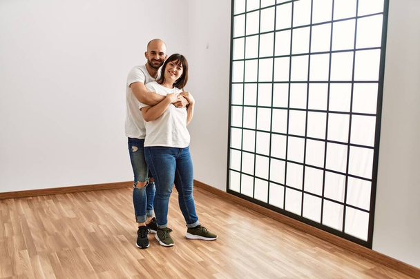Joven pareja hispana sonriendo feliz y abrazándose en un nuevo hogar vacío. - Foto, Imagen