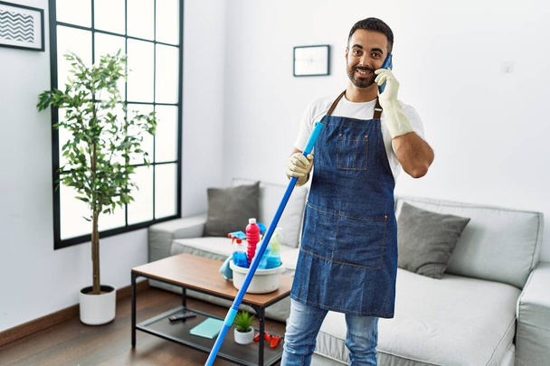 Young hispanic man wearing apron and gloves talking on the smartphone at home - Foto, immagini