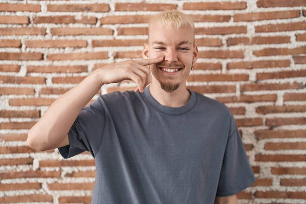 若い白人男性のレンガの壁の上に顔や鼻に手の指で指して立って、陽気な笑顔。美の概念  - 写真・画像