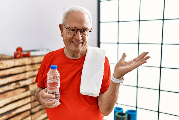 Senior man wearing sportswear and towel at the gym smiling cheerful presenting and pointing with palm of hand looking at the camera.  - Valokuva, kuva